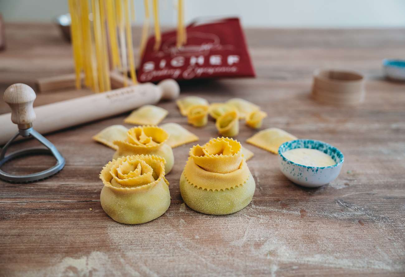 Handmade rose-shaped fresh pasta on a wooden table, with traditional Italian cooking tools and ingredients during a cooking class.